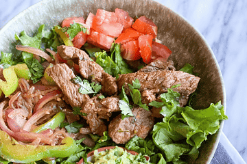 Steak fajitas in a bowl with lettuce, fajita veggies, guac