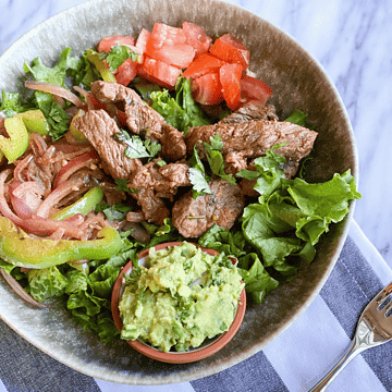 Steak fajitas in a bowl with lettuce, fajita veggies, guac